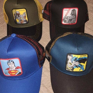 Könnte beinhalten: Vier Trucker-Hüte mit Aufnähern von Superhelden und Star Wars-Charakteren. Die Hüte haben Mesh-Rücken und gebogene Krempen. Die Aufnäher zeigen Boba Fett, Darth Vader, Superman und Batman. Die Hüte sind in verschiedenen Farben erhältlich.