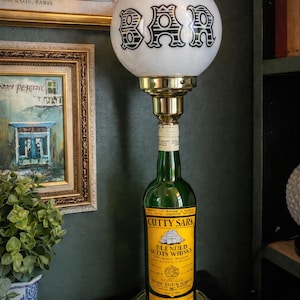 May include: A novelty lamp crafted from a green Cutty Sark whisky bottle. It has a white globe shade with the word "BAR" in black. The base and fittings are gold-toned, giving it a vintage appearance. The lamp is a decorative item.