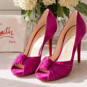May include: A pair of fuchsia suede peep-toe pumps with a knotted detail at the front, high stiletto heels, and signature red soles. The interior insoles feature the Christian Louboutin Paris logo, and a branded dust bag is visible in the background.