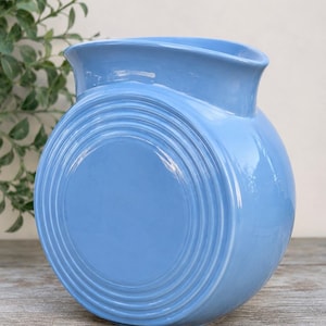 Puede incluir: Una jarra de cerámica azul claro con un cuerpo redondeado en forma de disco que presenta anillos circulares concéntricos en relieve en el lateral. El recipiente tiene un acabado liso y brillante, una boca ancha y un pico curvo.