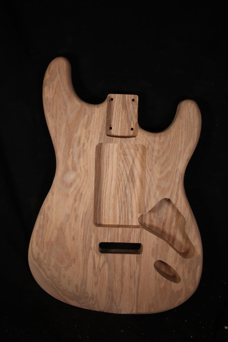 May include: Unfinished wooden guitar body with a natural wood grain finish. The body features cutouts for pickups, a tremolo system, and neck attachment. The guitar body is ready for customization and assembly.