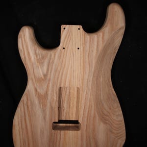 May include: Unfinished wooden guitar body with a natural wood grain pattern. The body has cutouts for the neck, pickups, and bridge. The wood has a light brown colour and a smooth texture, ready for customisation.