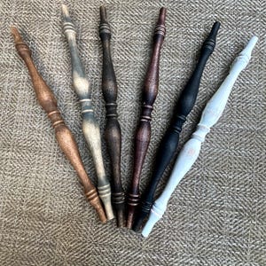 May include: Six slender, turned wooden spindles arranged in a fan shape. The spindles feature a decorative, elongated profile with multiple rounded sections and tapered ends, finished in various colors including light brown, grey, dark brown, and black.