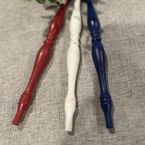 May include: Three turned wooden spindles arranged vertically, featuring a red spindle on the left, a white spindle in the center, and a blue spindle on the right. Each spindle has a smooth, tapered design with decorative ridges and a painted finish.