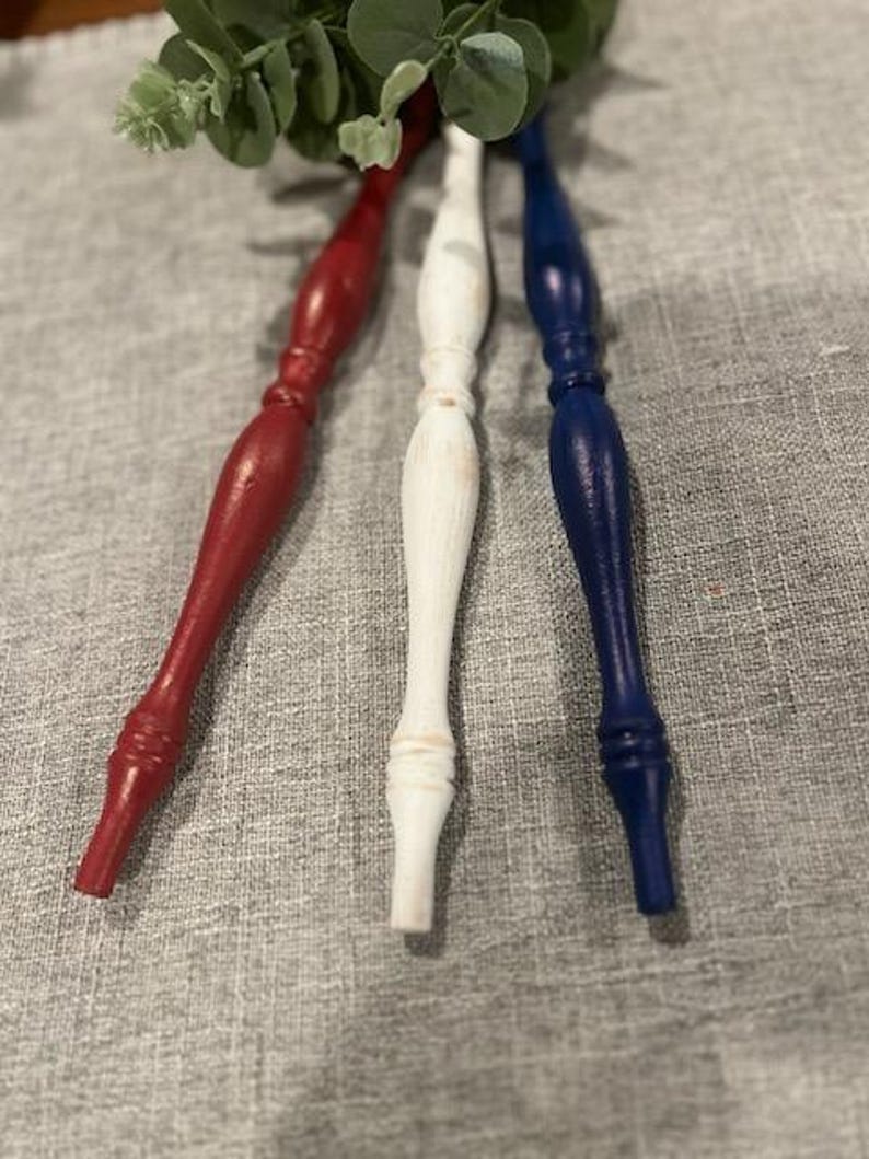 May include: Three turned wooden spindles arranged vertically, featuring a red spindle on the left, a white spindle in the center, and a blue spindle on the right. Each spindle has a smooth, tapered design with decorative ridges and a painted finish.