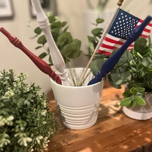 May include: A white ceramic pot with horizontal ridges containing three slender, tapered wooden sticks painted in solid red, white, and blue, alongside a small American flag on a wooden pole with a gold-colored finial tip.