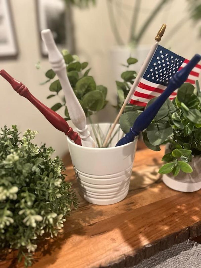 May include: A white ceramic pot with horizontal ridges containing three slender, tapered wooden sticks painted in solid red, white, and blue, alongside a small American flag on a wooden pole with a gold-colored finial tip.
