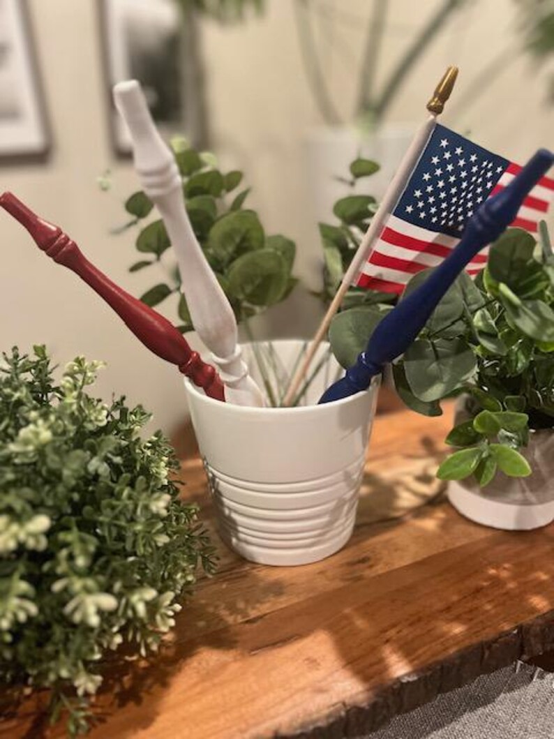 May include: A white ceramic cup containing three wooden wands painted in red, white, and blue, alongside a small American flag on a wooden pole with a gold finial. The wands feature a tapered, turned design with a smooth, matte finish.