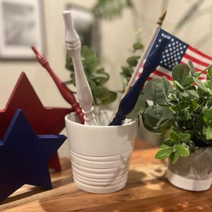 May include: A patriotic display with red, white, and blue wooden stars and decorative items. A small American flag and decorative wooden pieces are in a white ceramic pot. A small potted plant is also present.