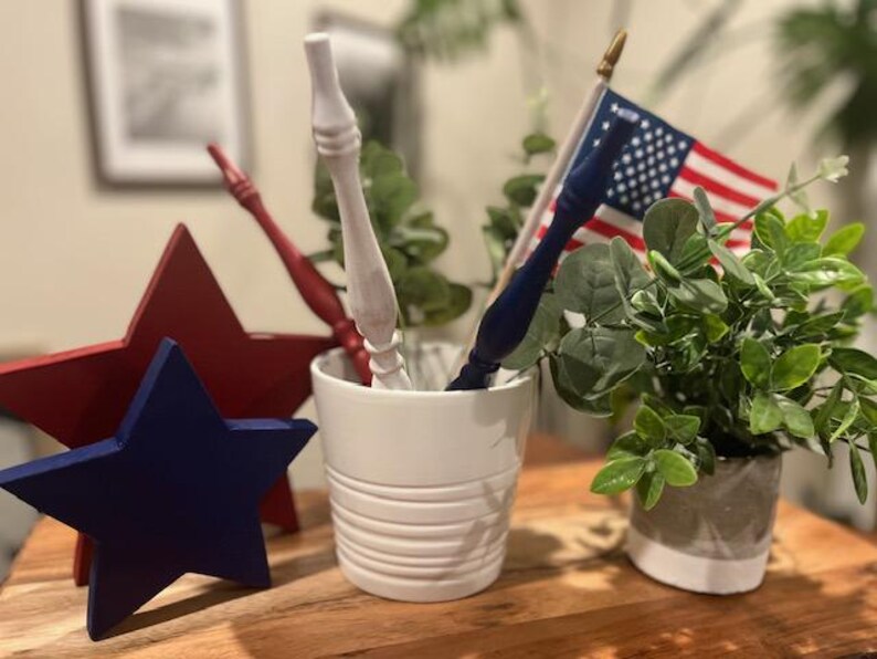 May include: A white ceramic pot containing a white wooden spindle, a red wooden spindle, a blue wooden spindle, and a small American flag. Beside the pot are a large red wooden star and a smaller blue wooden star, with a green potted plant.