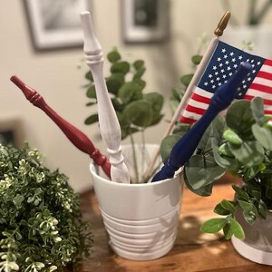 May include: A white ceramic cup containing three decorative wooden wands in red, white, and blue, alongside a small American flag on a wooden pole with a gold finial. The wands feature a turned, tapered design with a smooth, painted finish.