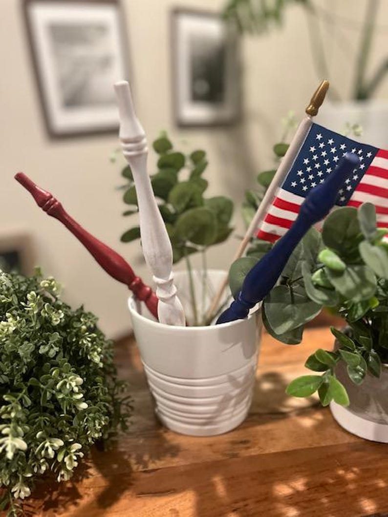 May include: A white ceramic cup containing three decorative wooden wands in red, white, and blue, alongside a small American flag on a wooden pole with a gold finial. The wands feature a turned, tapered design with a smooth, painted finish.