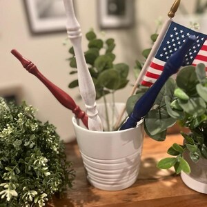May include: Three decorative wooden spindles in red, white, and blue are placed inside a white ceramic pot. A small American flag with red, white, and blue stripes and white stars is positioned behind the blue spindle, featuring a gold-colored finial.