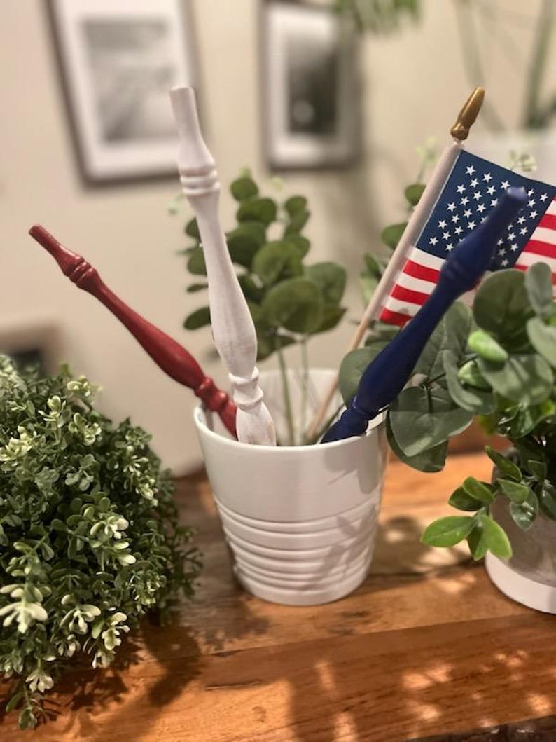 May include: Three decorative wooden spindles in red, white, and blue are placed inside a white ceramic pot. A small American flag with red, white, and blue stripes and white stars is positioned behind the blue spindle, featuring a gold-colored finial.