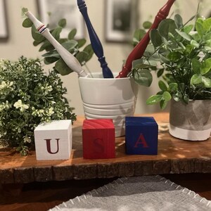 May include: Three wooden blocks painted white, red, and blue, each featuring a single letter to spell USA. The white block has a red U, the red block has a blue S, and the blue block has a red A. Behind them are three turned wooden spindles.