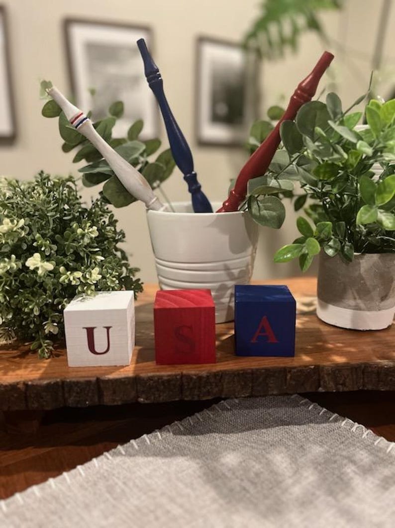 May include: Three wooden blocks painted white, red, and blue, each featuring a single letter to spell USA. The white block has a red U, the red block has a blue S, and the blue block has a red A. Behind them are three turned wooden spindles.