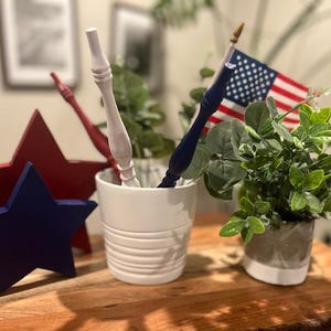 May include: A white ceramic pot containing a white wooden spindle, a blue wooden spindle, and a small American flag. Beside the pot are a large red wooden star and a smaller blue wooden star, along with a small potted green plant in a gray pot.