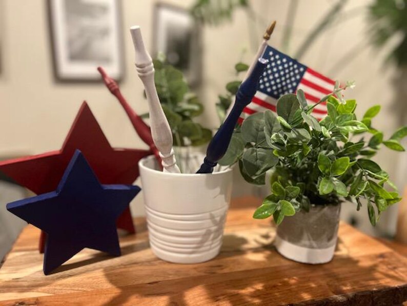 May include: A white ceramic pot containing a white wooden spindle, a blue wooden spindle, and a small American flag. Beside the pot are a large red wooden star and a smaller blue wooden star, along with a small potted green plant in a gray pot.