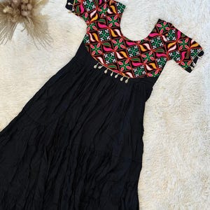 May include: A black maxi dress with a colorful embroidered bodice. The bodice features a geometric pattern in shades of pink, green, yellow, and white. The dress has a tiered skirt and is adorned with silver beads.