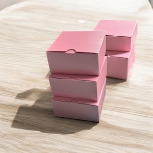 May include: Four pink square boxes with tab closures are stacked. The boxes are arranged in a staggered formation. The boxes are made of a matte paper material. The boxes are a soft, pastel pink colour.