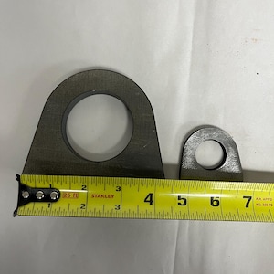 May include: Two dark gray metal mounting tabs with circular holes, positioned next to a yellow Stanley measuring tape. The larger tab measures approximately 3.5 inches in width, while the smaller tab measures approximately 1.5 inches in width.