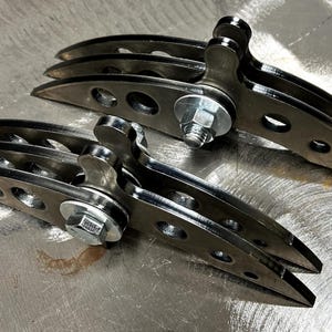 May include: Two identical metal mechanical assemblies, each featuring three curved, dark-finished steel plates with circular cutouts. The plates are joined by a central silver-colored bolt, a nut, and a washer, creating a layered, arched structure.