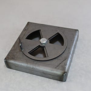 May include: A square, raw steel metal plate featuring a circular, four-spoke rotating vent cover secured in the center by a single silver-colored hexagonal bolt. The metal surface displays a dark, industrial finish with visible weld marks.