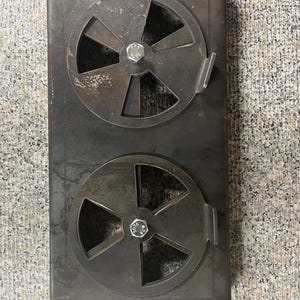 May include: A rectangular, dark metal stove door with two circular, wheel-shaped vents. Each vent has a central bolt and a handle. The metal has a distressed appearance, with visible wear and tear.