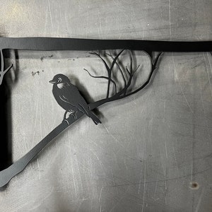 May include: Black metal wall-mounted shelf bracket with a bird and branch design. The bracket has a hook on the end.
