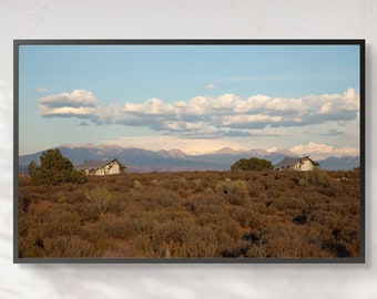 May include: A photograph of a landscape with two small white houses in the foreground, a field of brown brush, and a mountain range in the background. The sky is blue with white clouds.