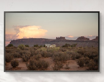 May include: A framed landscape photograph featuring a desert scene with a small building and distant mesas under a sunset sky. The foreground is filled with desert vegetation, and the frame is black.