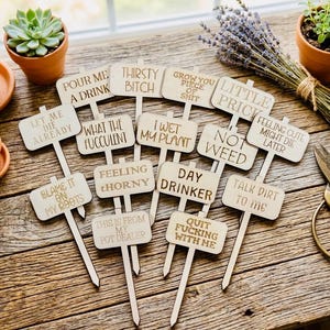 May include: A collection of wooden plant markers with humorous phrases like "Pour Me A Drink" and "Thirsty Bitch" laser-etched on them. The markers are light brown and have a stake for easy insertion into soil. The image includes potted plants and gardening tools.
