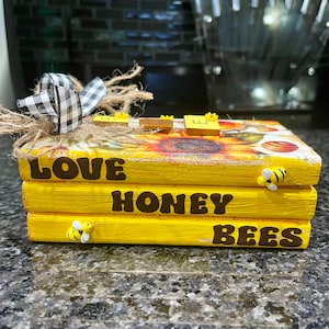 May include: A yellow wooden box with the words "Love Honey Bees" painted on it. The box is decorated with a sunflower and a black and white checkered ribbon. There are small bee figurines on the box.