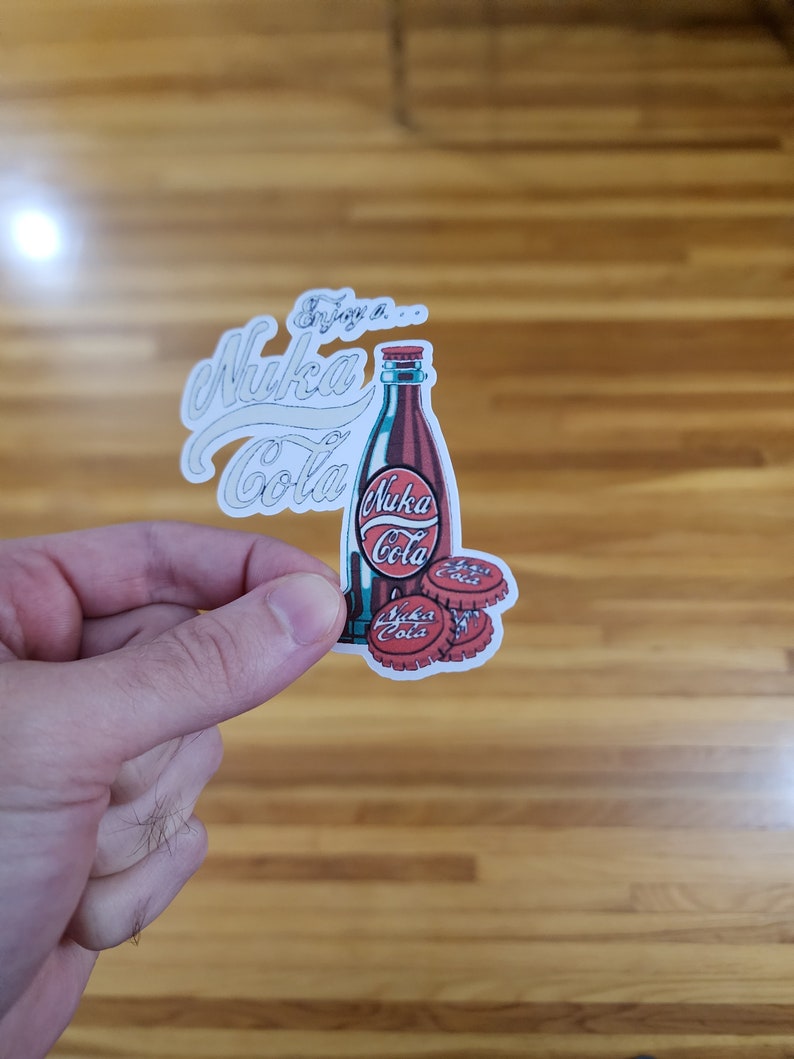 May include: A white sticker with a red and blue illustration of a bottle of Nuka Cola. The bottle has a red cap and the text "Enjoy a... Nuka Cola" is written in white cursive above the bottle. Three bottle caps are below the bottle, with the text "Nuka Cola" on each.
