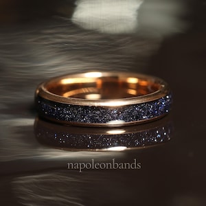 May include: A rose gold band ring with a dark blue glitter inlay. The ring is on a dark reflective surface.