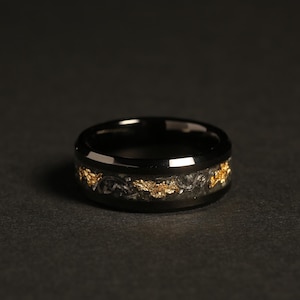 May include: A black ring with a band of gold leaf and dark gray material. The ring has a polished, reflective surface and a dark background. The gold leaf is irregularly shaped and adds a decorative element to the ring.