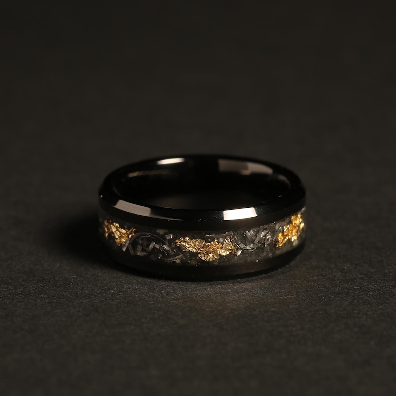 May include: A black ring with a band of gold leaf and dark gray material. The ring has a polished, reflective surface and a dark background. The gold leaf is irregularly shaped and adds a decorative element to the ring.