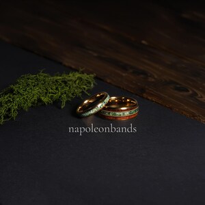 May include: Two gold wedding rings with unique designs. One ring features a band of green stone, while the other has a wood grain and green stone inlay. The rings are displayed on a black surface with the text "napoleonbands".