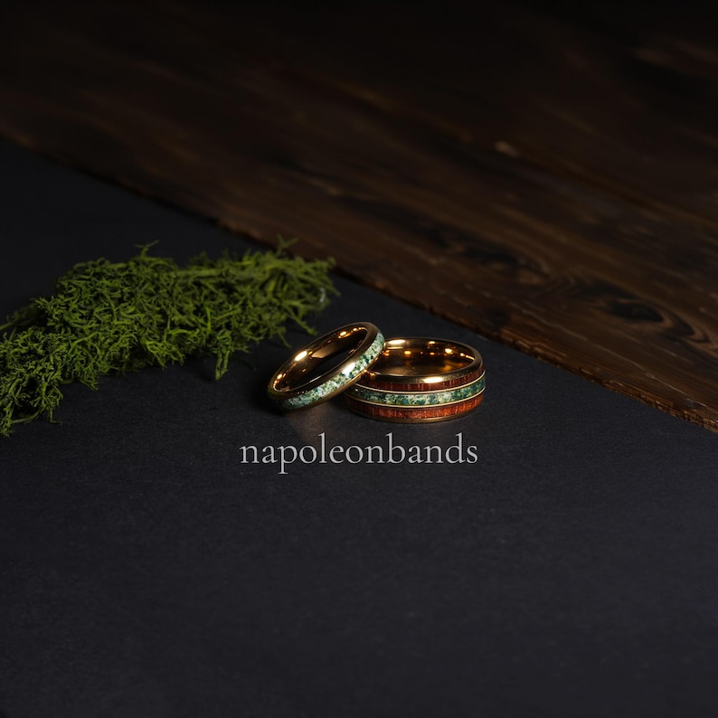 May include: Two gold wedding rings with unique designs. One ring features a band of green stone, while the other has a wood grain and green stone inlay. The rings are displayed on a black surface with the text "napoleonbands".