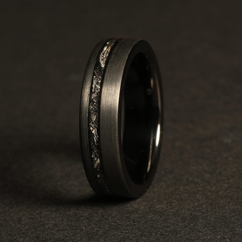 May include: A black tungsten carbide ring with a silver inlay. The ring has a brushed finish and a beveled edge.