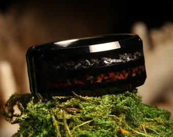 May include: A black tungsten carbide ring with a thin band of red and black inlay. The ring is sitting on a bed of green moss.