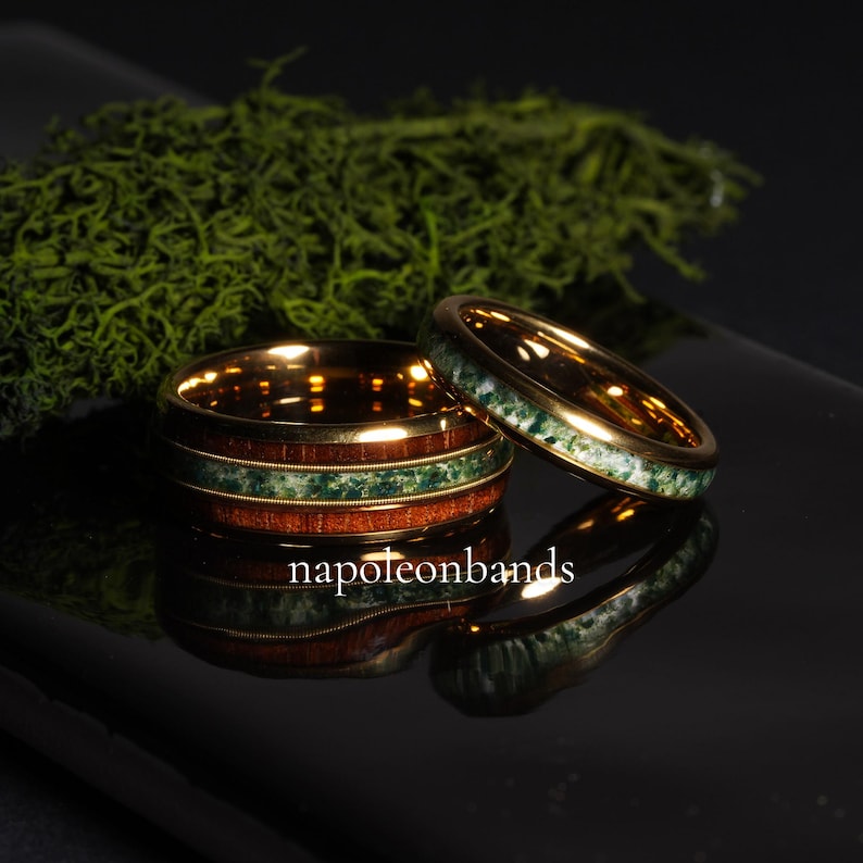 May include: Two gold wedding rings with a wood and green stone inlay. The rings are stacked on a reflective black surface, with the text "napoleonbands" visible. Green moss is in the background.