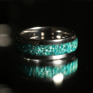 May include: A silver-colored ring with a band of teal-colored, crushed glass-like material. The ring is highly reflective, with a dark background. The ring is a wide band style.