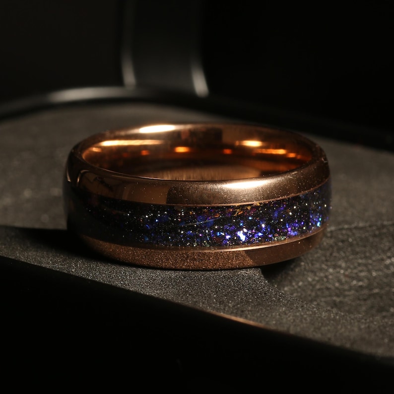May include: A rose gold ring with a dark band inlaid with sparkling blue and purple flecks. The ring has a polished finish and a classic, rounded design. The band is approximately 0.25 inches wide.
