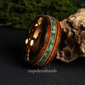 May include: A gold-colored ring with a wood and stone inlay. The ring features a band of reddish-brown wood, a band of green stone, and a thin gold band. The text "napoleonbands" is at the bottom of the image.
