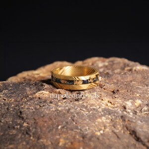May include: A gold ring with a textured pattern and a black and gold inlay. The ring is displayed on a rough, brown stone. The text "napoleonbands" is visible at the bottom.
