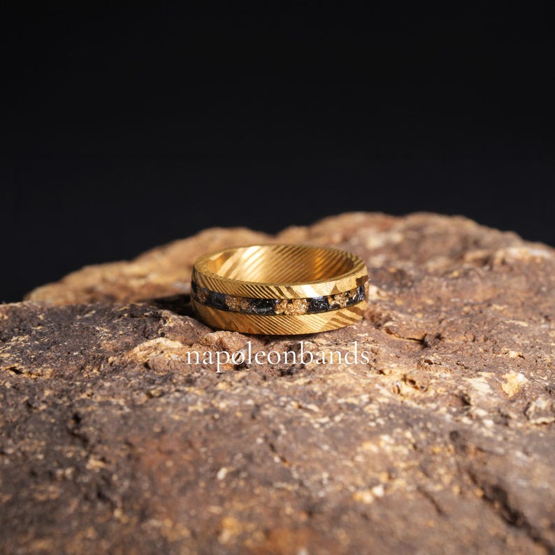 May include: A gold ring with a textured pattern and a black and gold inlay. The ring is displayed on a rough, brown stone. The text "napoleonbands" is visible at the bottom.