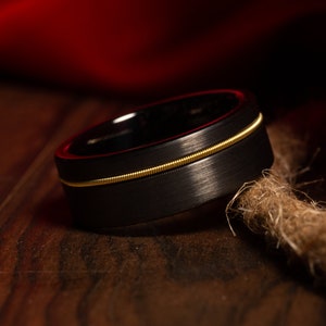 May include: A black ring with a gold-coloured string inlay. The ring has a brushed metal finish and a sleek, modern design. The gold string adds a unique touch. The ring is a piece of jewellery. The ring's diameter is approximately 2 cm.