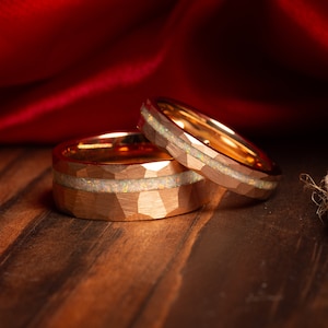 May include: Two gold wedding bands with a hammered finish and a white opal inlay. The bands are stacked on top of each other on a wooden surface.