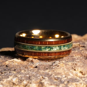 May include: A gold-colored ring with a band of dark wood, a central stripe of green stone, and thin gold bands. The ring is set on a textured, brown surface. The text "napoleonbands" is visible.